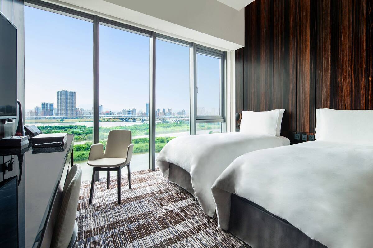 A hotel room with two beds, a desk, a chair, and a large window offering a city view.