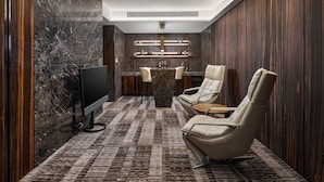 A modern, well-lit room with a marble wall, a flat-screen TV, a marble coffee table, two leather armchairs, and a wooden sideboard with shelves.