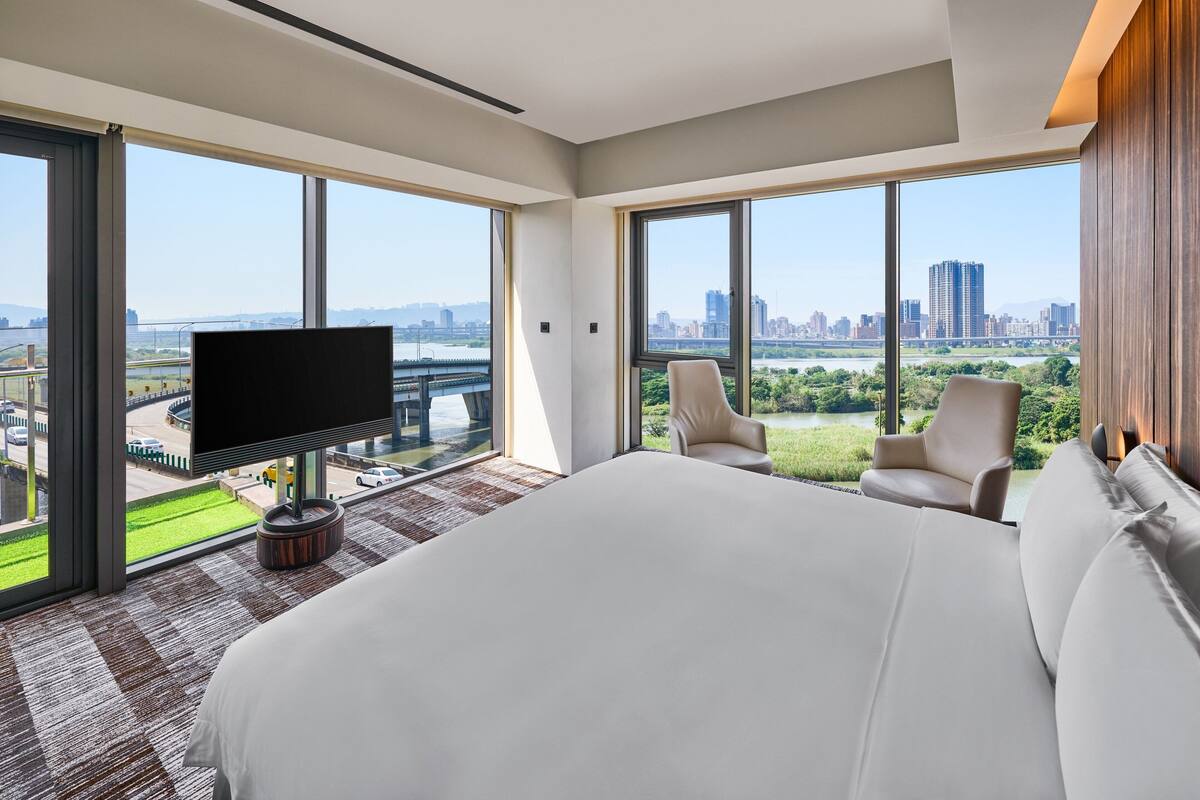 A modern hotel room with a large bed, a TV, and a view of a cityscape and greenery.