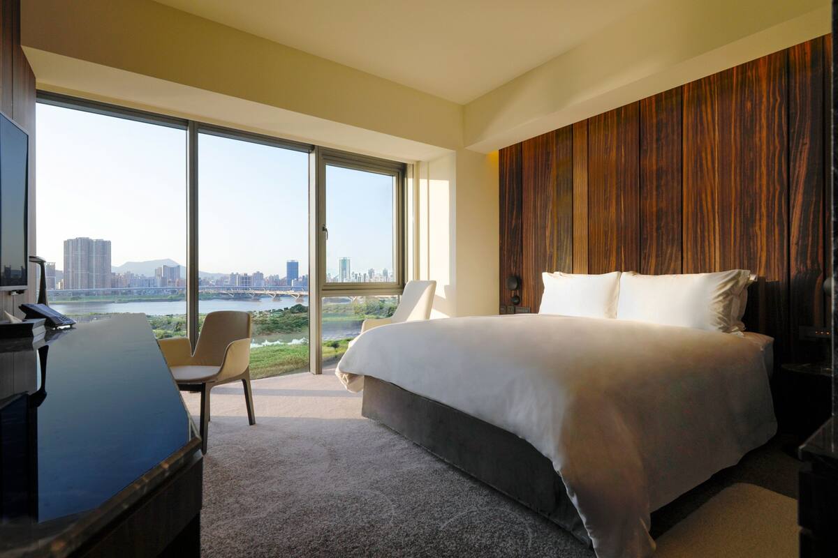 A modern hotel room with a large bed, a desk, and a chair. The room has a large window offering a view of a cityscape and a river.