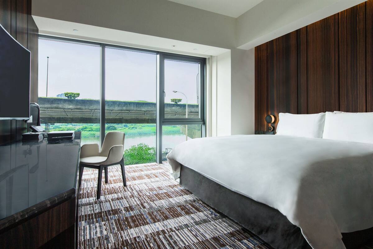 A modern hotel room with a large bed, a desk with a chair, and a view of a green landscape through large windows.