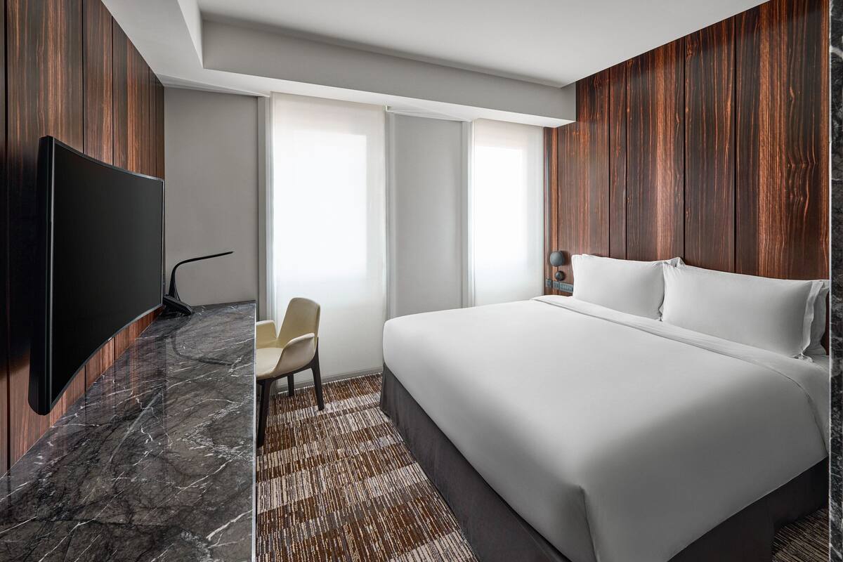 A modern hotel room with a large bed, a flat-screen TV mounted on the wall, a small chair, and a window with curtains.