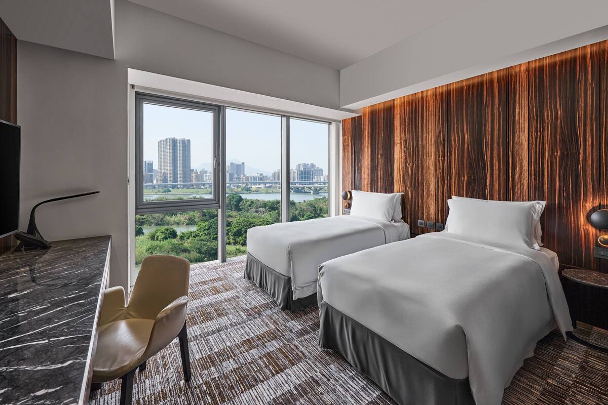 A hotel room with two beds, a desk, a chair, and a large window offering a city view.