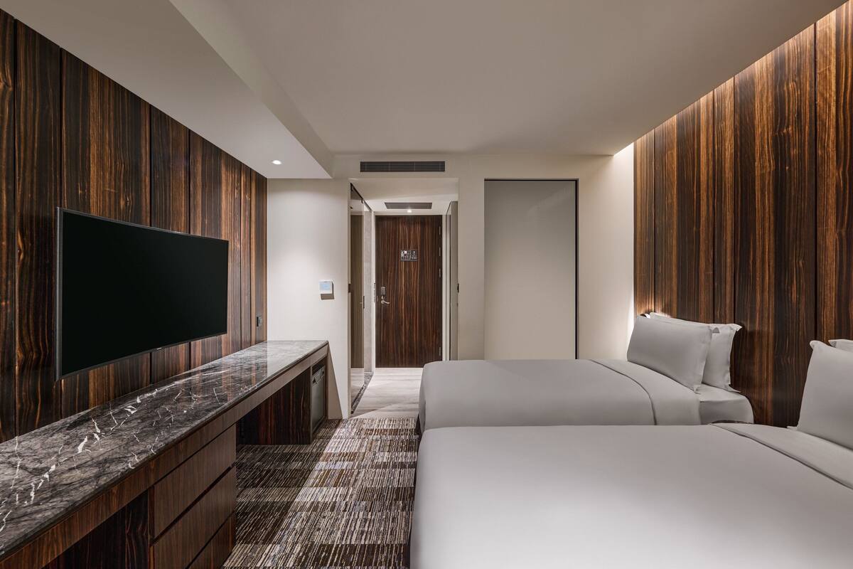 A modern hotel room with a large flat-screen TV, a marble countertop, a bed with white linens, and wooden panel walls.