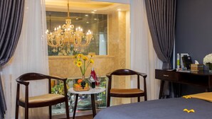 Quarto de hotel com uma cama grande, duas cadeiras de madeira, uma mesinha redonda com uma cesta de frutas e copos de vinho, uma escrivaninha com abajur, e vista da cidade através de uma janela ampla com cortinas.