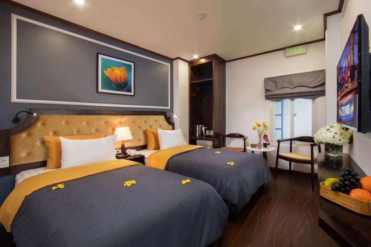 Habitación de hotel con dos camas, un cabecero grande, un televisor, una pequeña mesa con flores y una mesita de noche de madera con fruta.