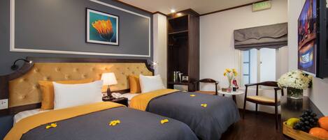 A hotel room with two beds, a large headboard, a TV, a small table with flowers, and a wooden side table with fruit.