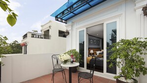 Uma varanda com mesa e cadeiras, um vaso de planta e vista do interior através de uma porta de vidro.