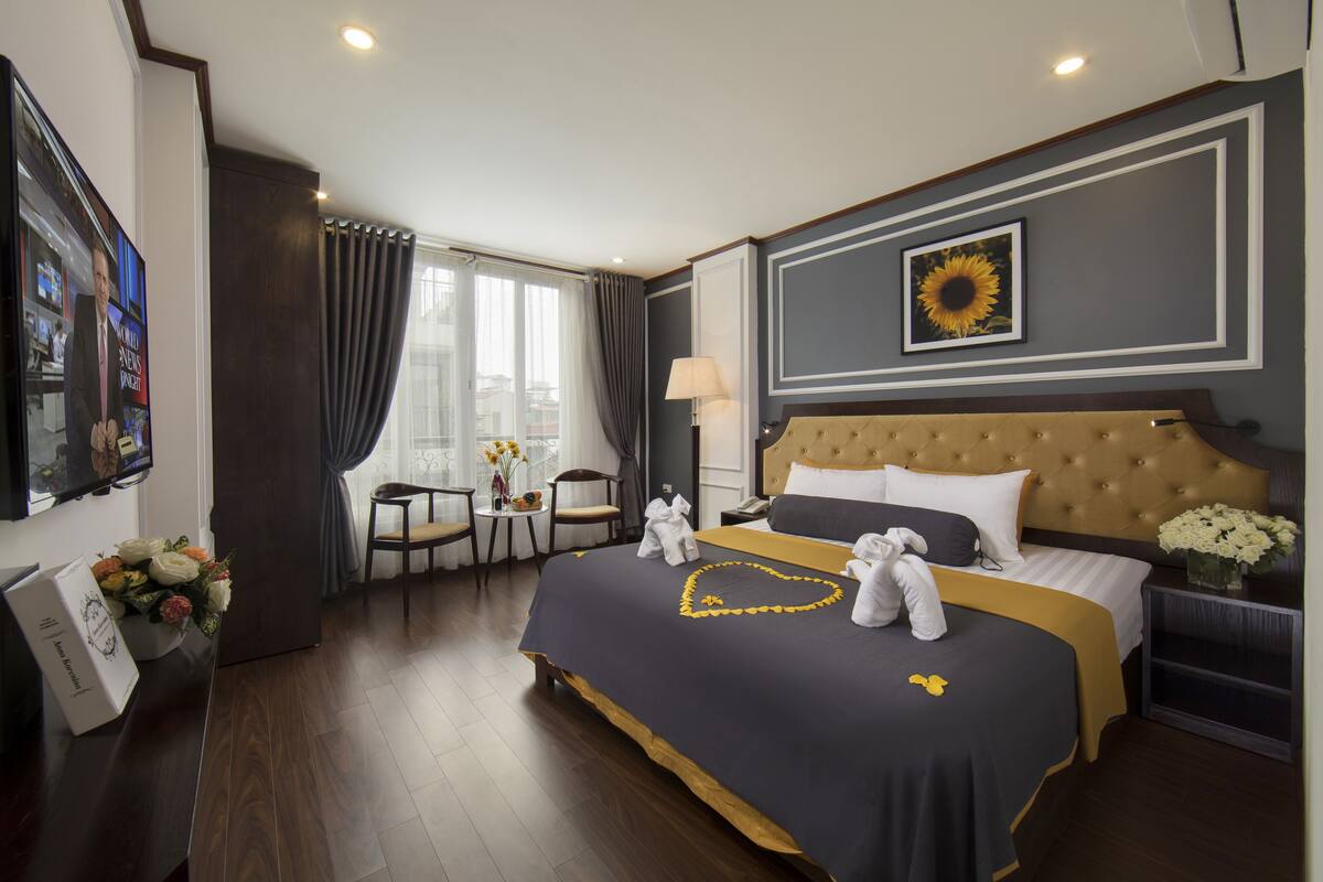 Habitación de hotel con una cama grande, cabecera, mesita de noche, lámpara, jarra con flores, una silla, una mesa con un jarrón de flores y un televisor en la pared.