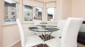 Une pièce d’angle avec une table en verre et des chaises blanches, de grandes fenêtres et une vue sur les immeubles voisins.