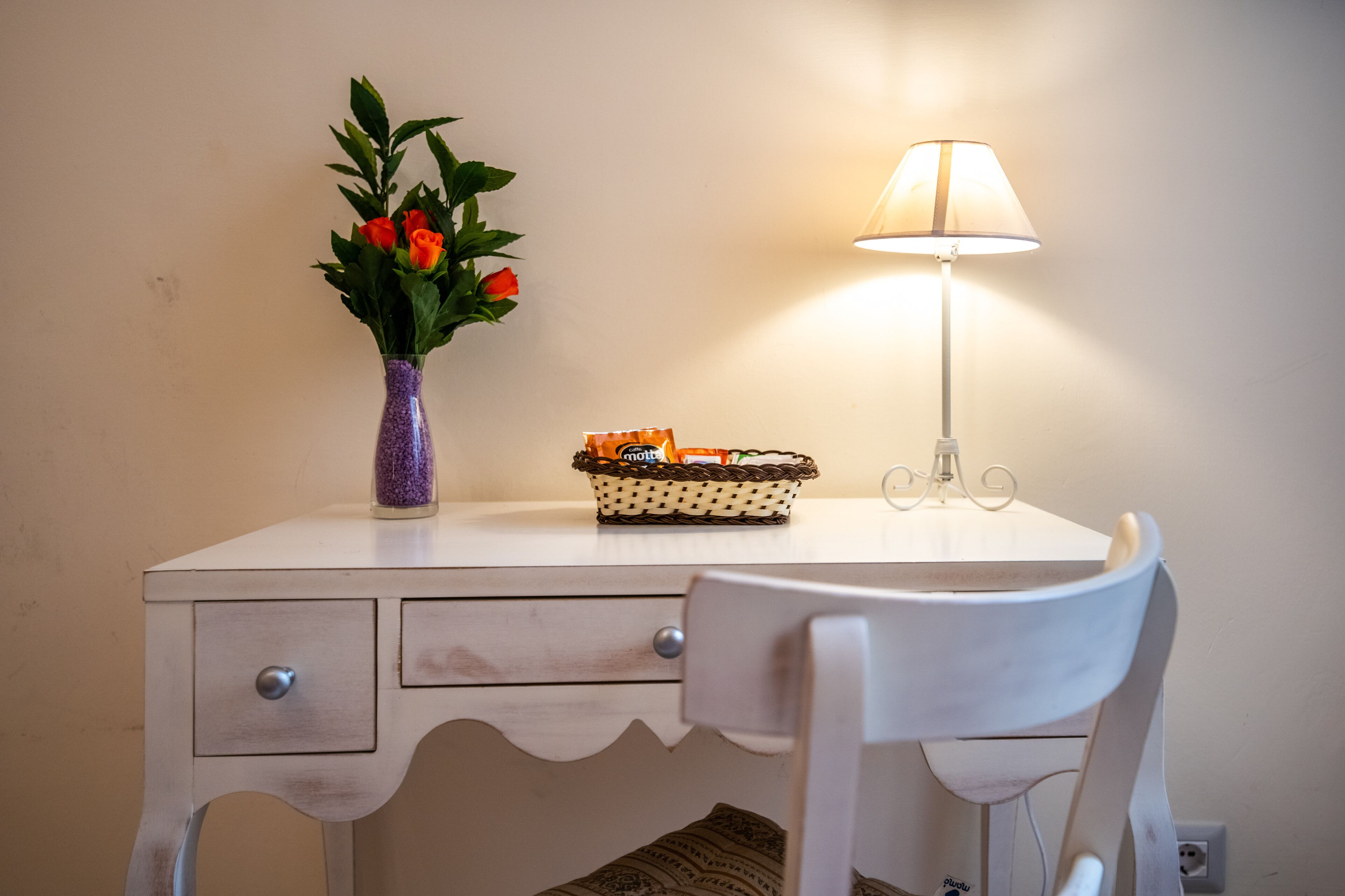 Un escritorio blanco con un jarrón morado de flores, una cesta de naranjas y una lámpara de mesa.