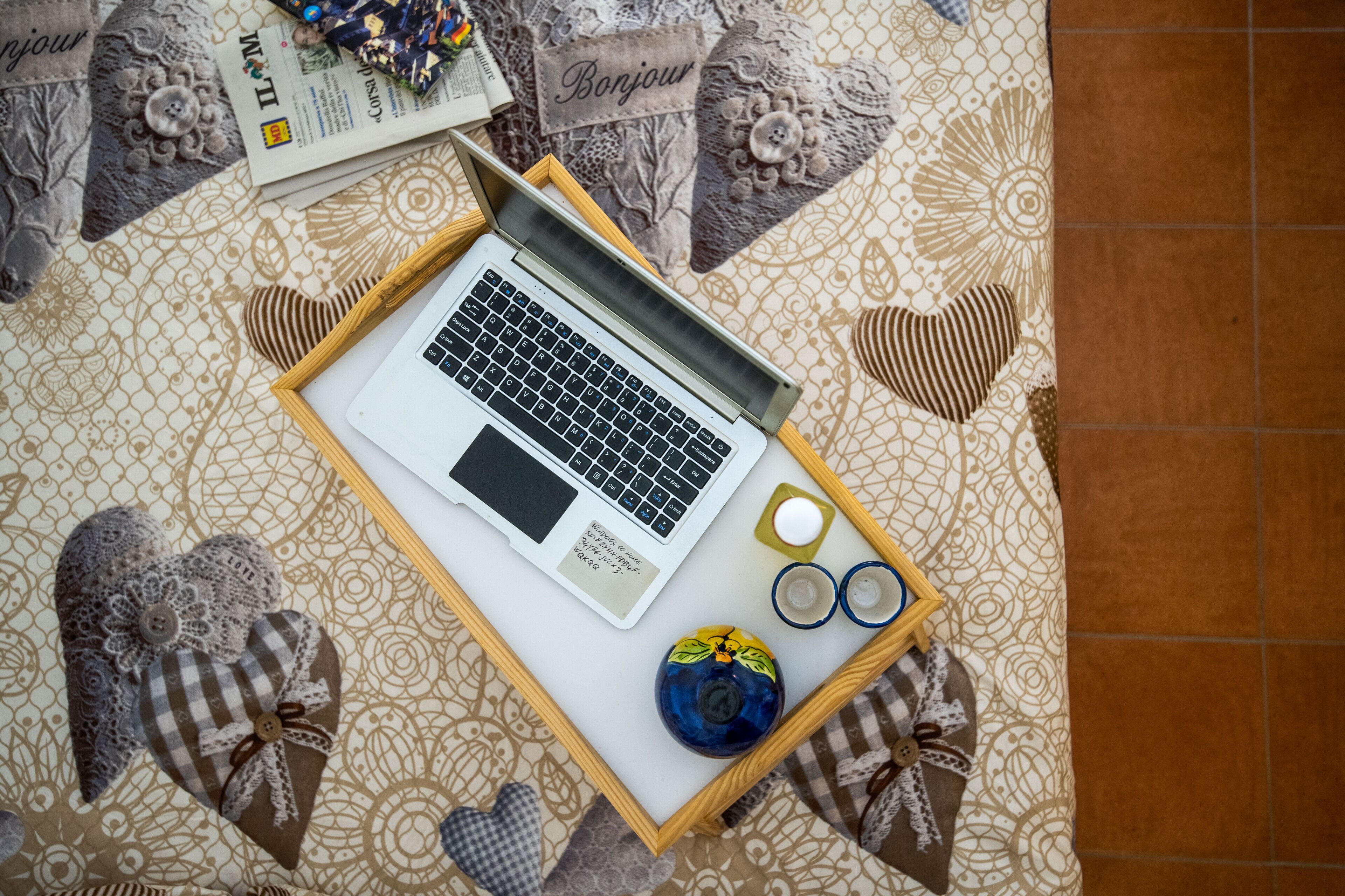 Ein Laptop auf einem Tablett mit einem dekorativen herzförmigen Kissen, einer kleinen blauen Vase und einer Brille.