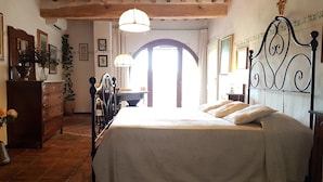 Un dormitorio con cama, cómoda de madera y escritorio con silla. Se ve una ventana con cortinas, un candelabro y una cruz en la pared.
