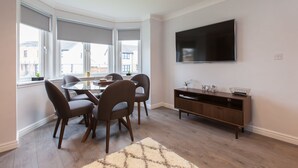A modern living room with a dining table, chairs, a flat-screen TV, and a large window with blinds.
