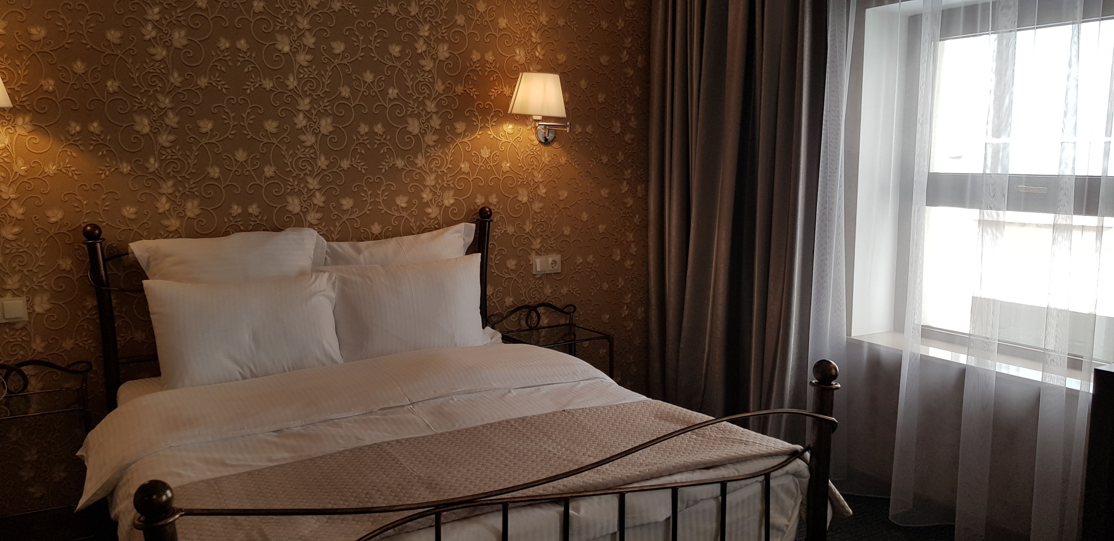 Un lit bien fait, avec du linge de lit blanc, dans une pièce dont les murs sont recouverts d’un papier peint à motifs et qui possède une fenêtre ornée de rideaux légers.