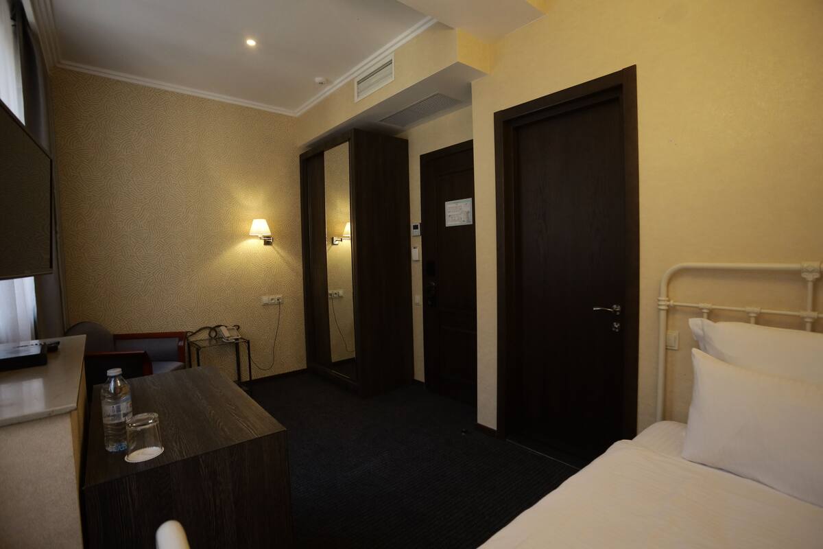 A hotel room with a bed, a desk with a lamp, a chair, and a small table with a bottle and glass.