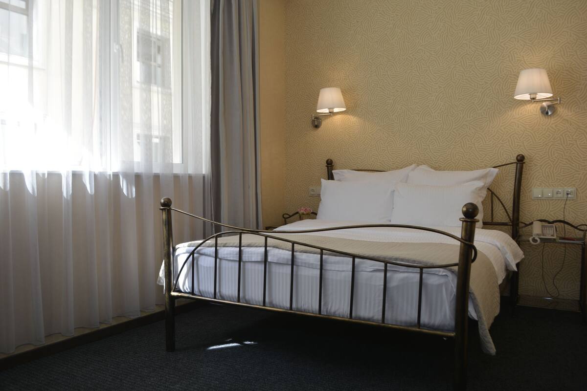 A hotel room with a metal-framed bed, two wall-mounted lamps, and a window with sheer curtains.