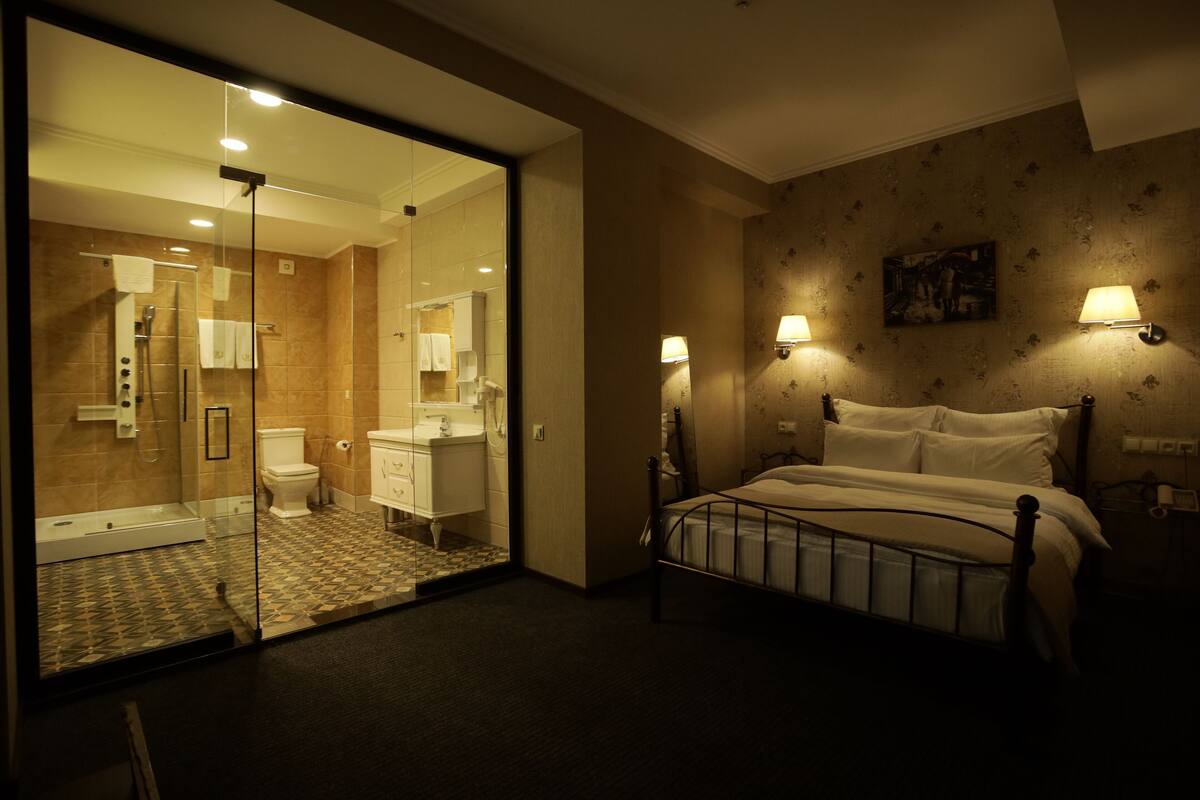 A hotel room with a glass shower enclosure, a bed with a metal frame, and two wall-mounted lamps.