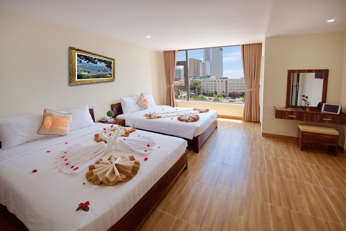 A hotel room with two beds, a large window with a city view, a desk with a mirror, and a framed picture on the wall.
