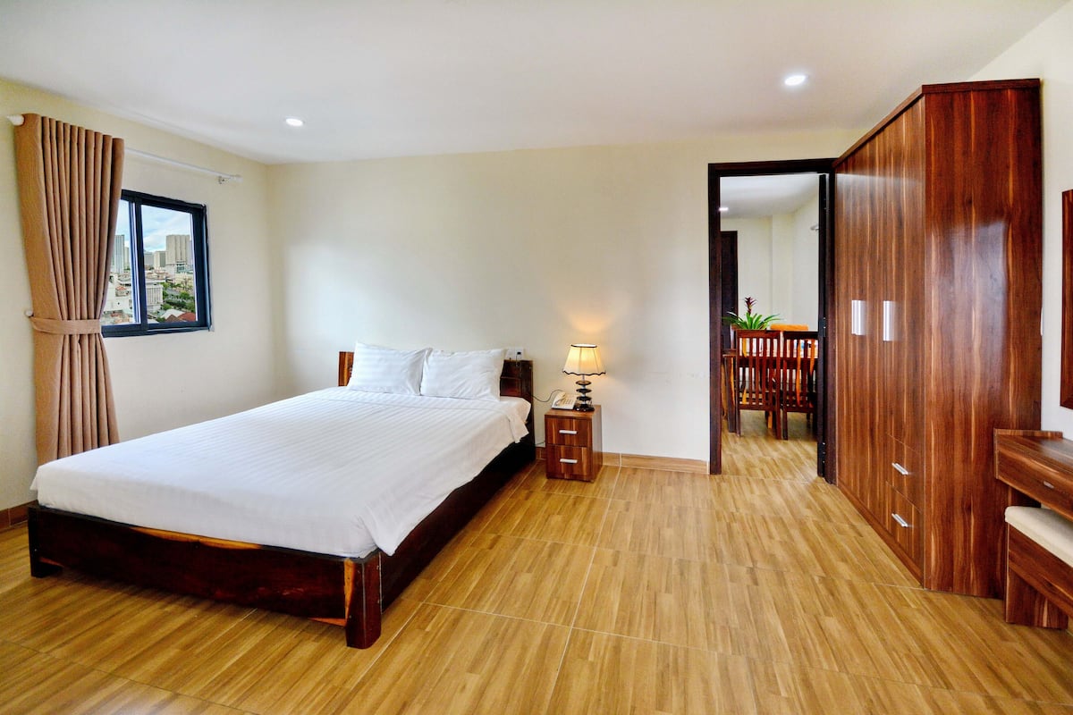 A hotel room with a large bed, wooden floor, a nightstand with a lamp, a wardrobe, and a view of the city through a window.