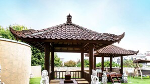 A gazebo with a tiled roof, a wooden bench, and a swing set in a park.