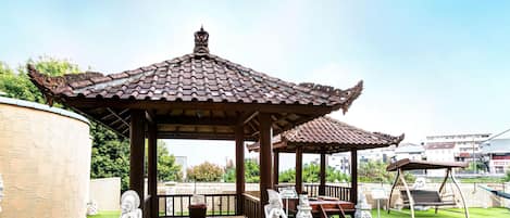 A gazebo with a tiled roof, a wooden bench, and a swing set in a park.