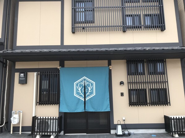 A building facade with a blue banner featuring a white logo, black-framed windows, and a tiled entrance area. - Guest House One More Heart at NARA DEN - Hostel (Nara)