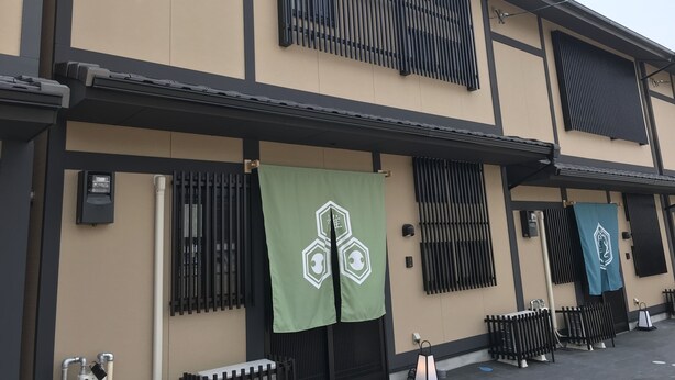 A building with a beige facade, black window frames, and green banners with white symbols. - Guest House One More Heart at NARA DEN - Hostel (Nara)