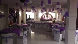 Un salón de banquetes con mesas redondas cubiertas con manteles blancos y cintas moradas, decorado con globos morados y blancos.