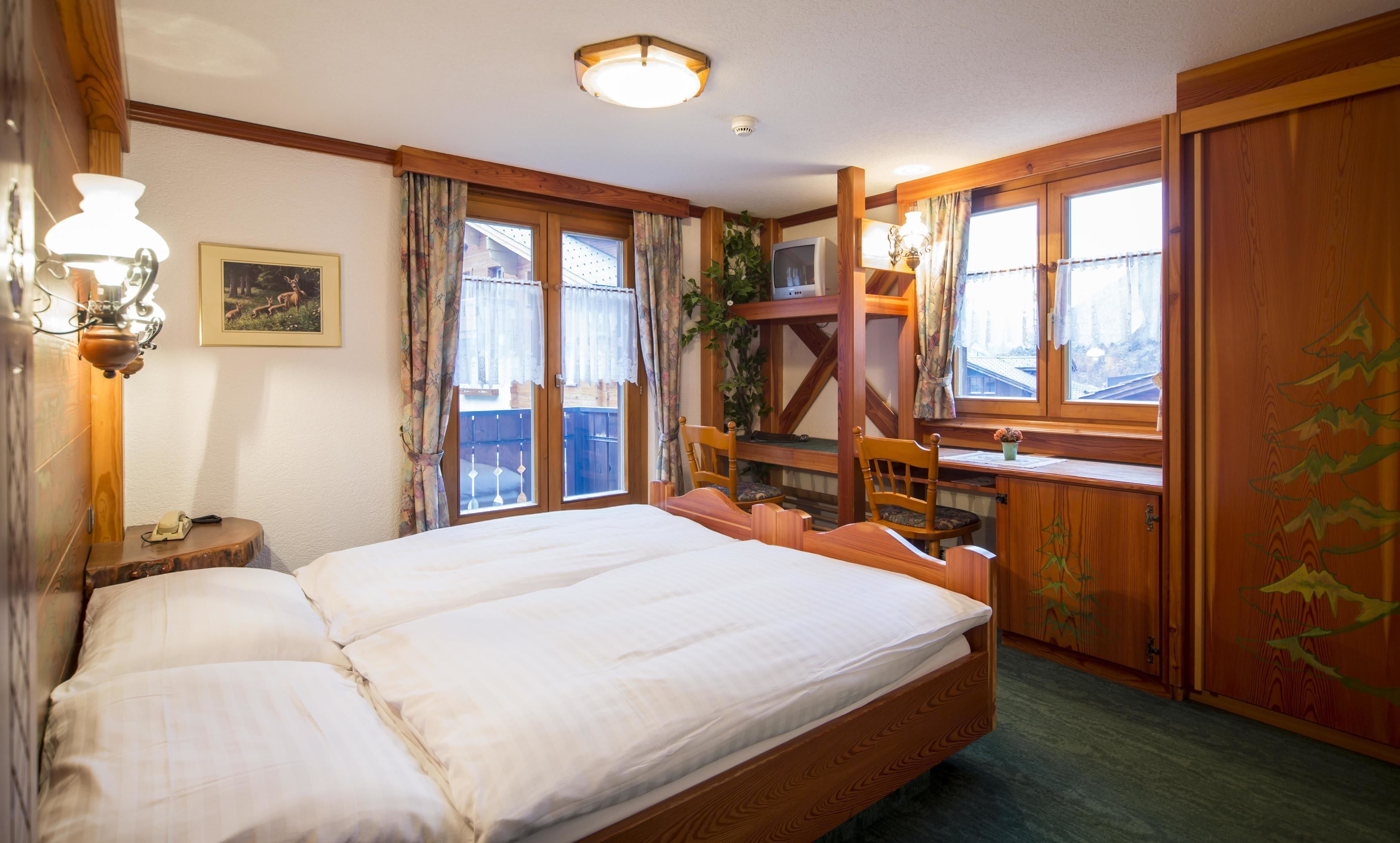 Ein Doppelbett mit Holz Kopfteil, ein Nachttisch mit Lampe, ein Fernseher und ein Fenster mit Vorhängen.
