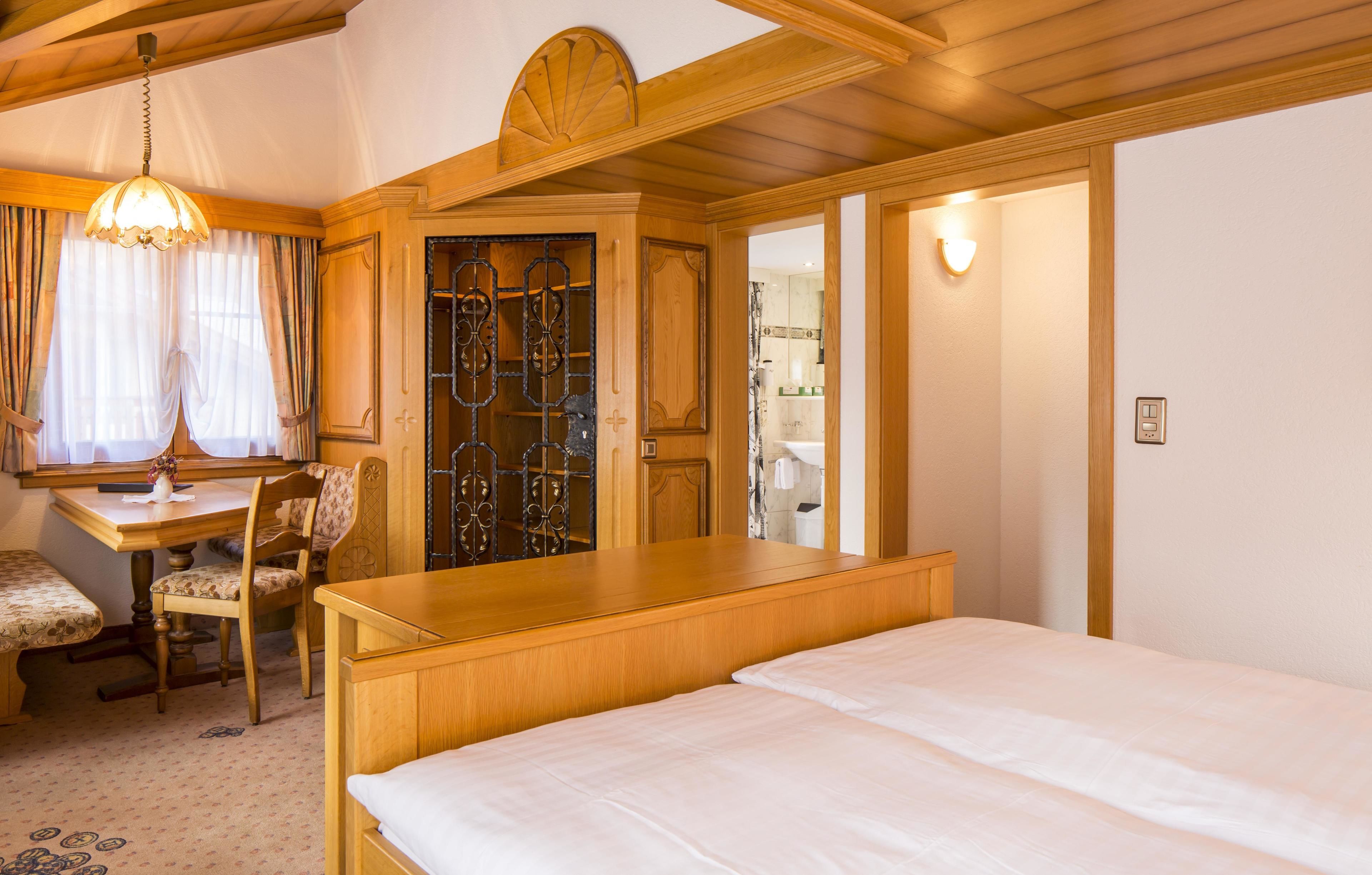 A hotel room with a wooden bed, a desk with a chair, a dining table with a bench, and a large window with curtains.