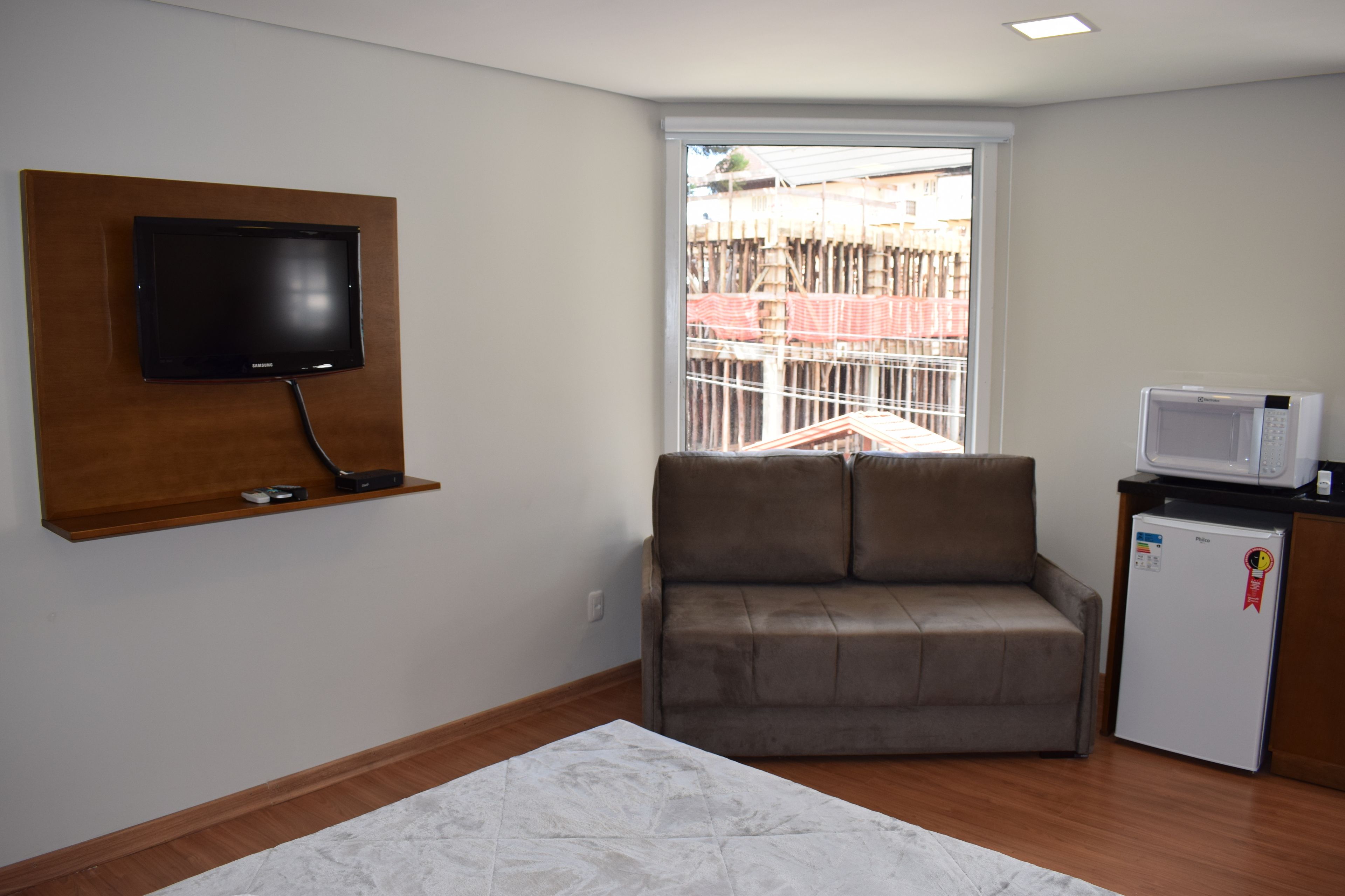 Una sala pequeña con una televisión de pantalla plana montada en un panel de madera, un sofá marrón, un refrigerador blanco y una ventana con vista a una obra en construcción.