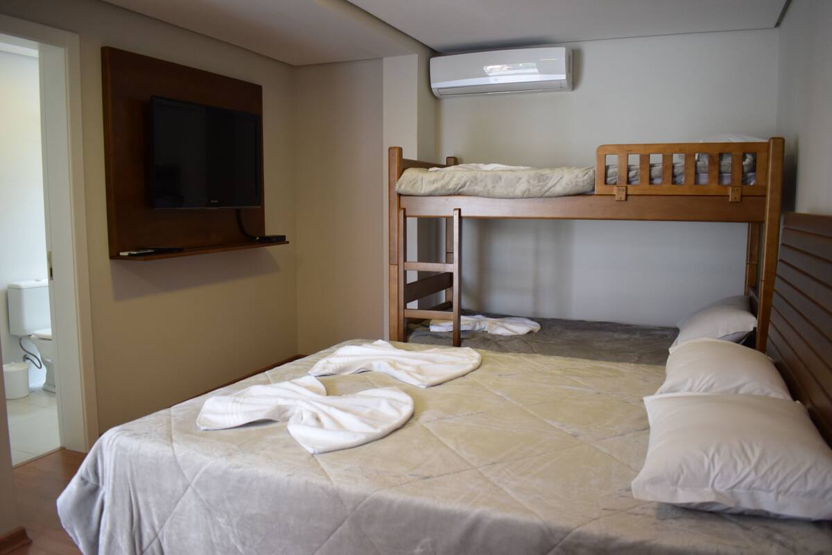 Une chambre avec un lit superposé, un téléviseur fixé au mur et une salle de bain visible à travers une porte ouverte.