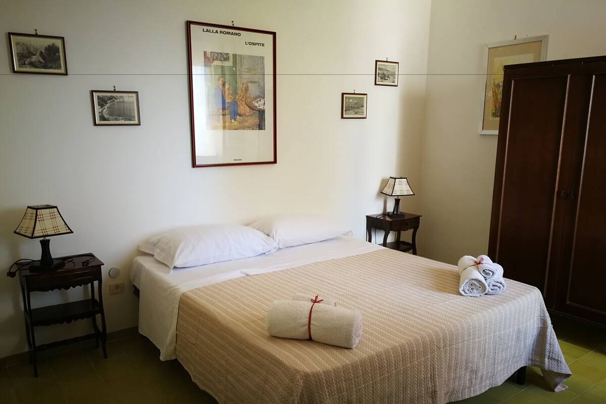 Un lit bien fait, avec une couette blanche et des oreillers, une table de chevet avec une lampe, une serviette roulée posée sur le lit et des tableaux encadrés accrochés au mur.