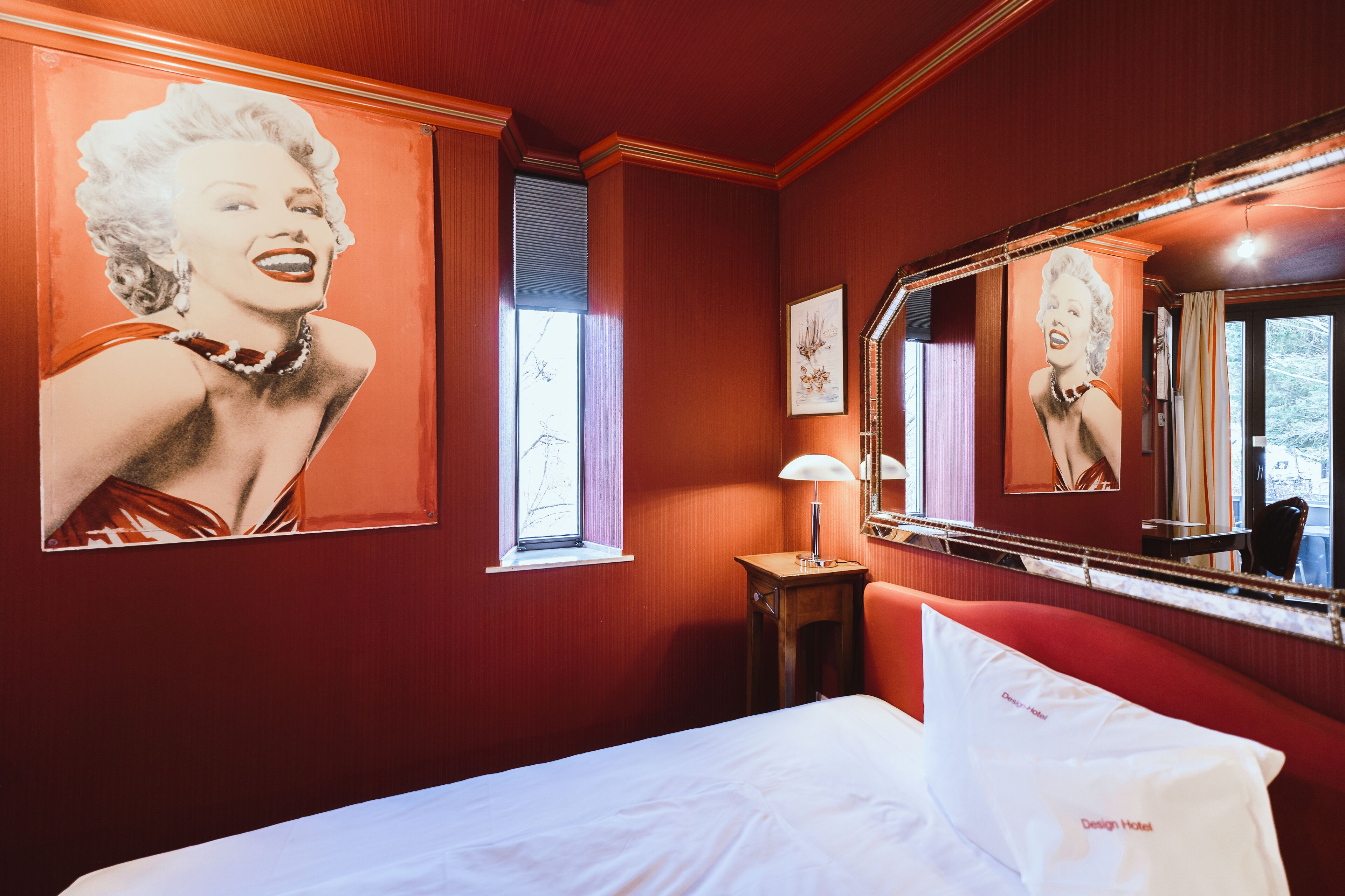 A hotel room with a large poster of Marilyn Monroe, a bed with white linens, a nightstand with a lamp, and a mirror reflecting the poster.