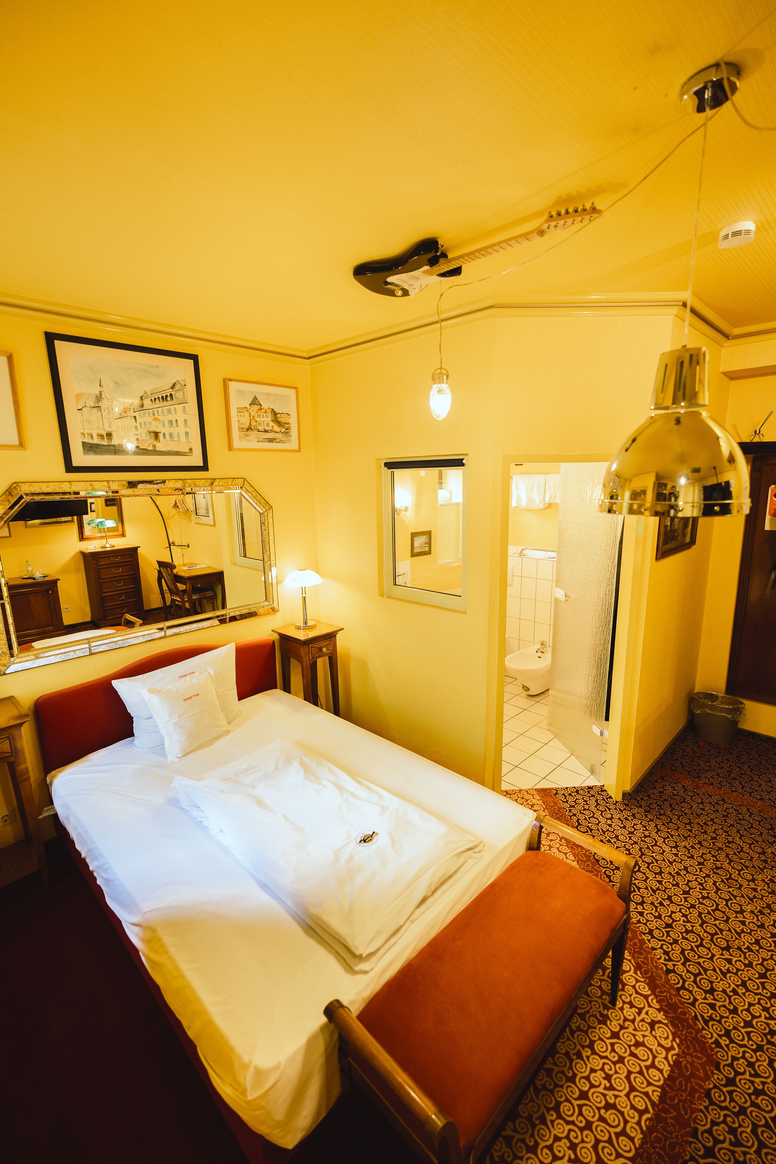 Habitación de hotel con cama, mesita de noche, espejo, lámpara, puerta de baño y suelo estampado.
