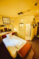 Habitación de hotel con cama, mesita de noche, espejo, lámpara, puerta de baño y suelo estampado.