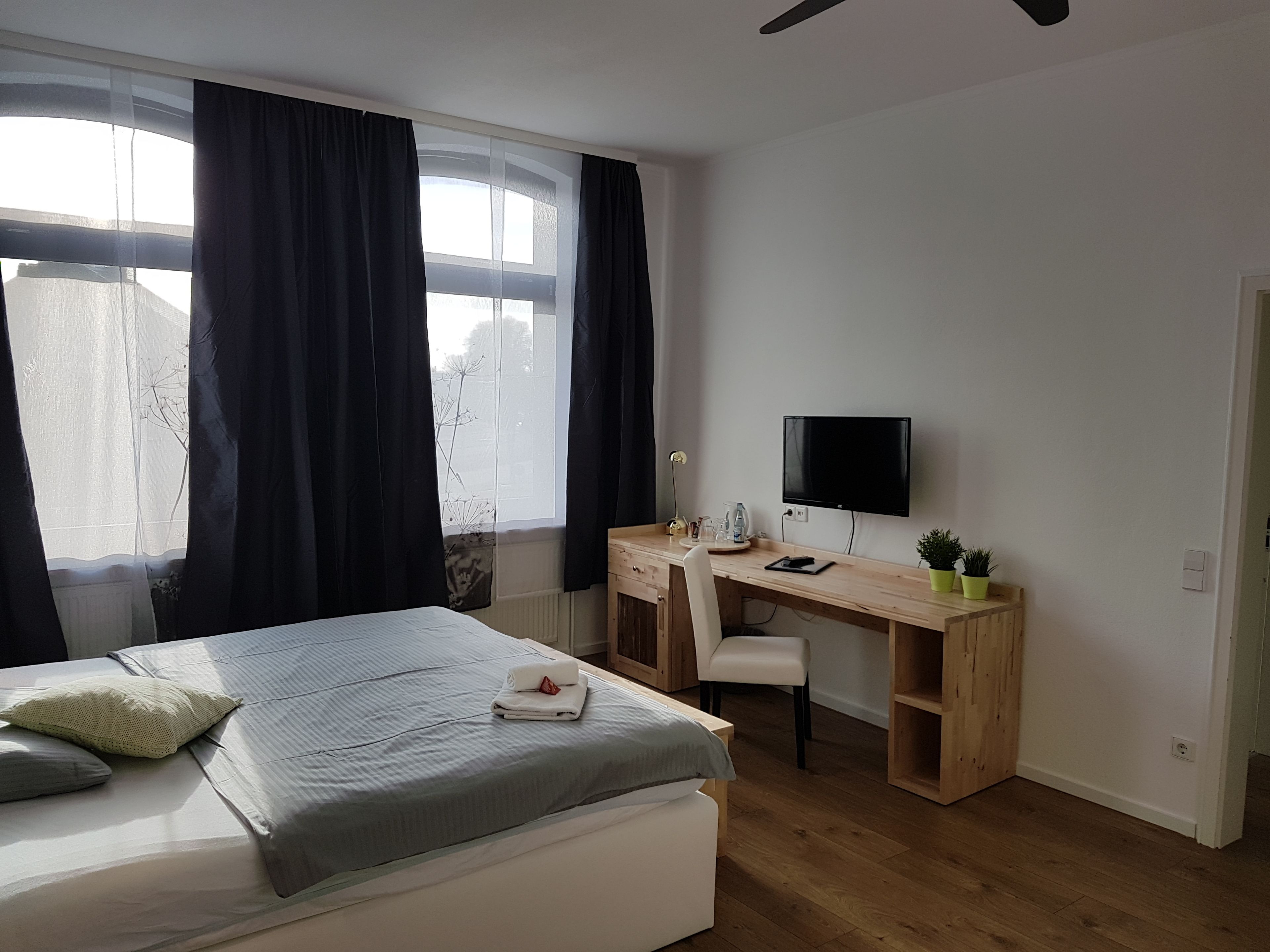 A bedroom with a bed, a desk, a chair, a TV, and a window with curtains.