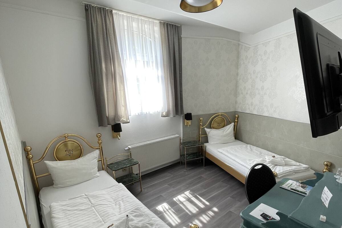 Habitación de hotel con dos camas individuales, un escritorio, una silla, un televisor y una ventana con cortinas.