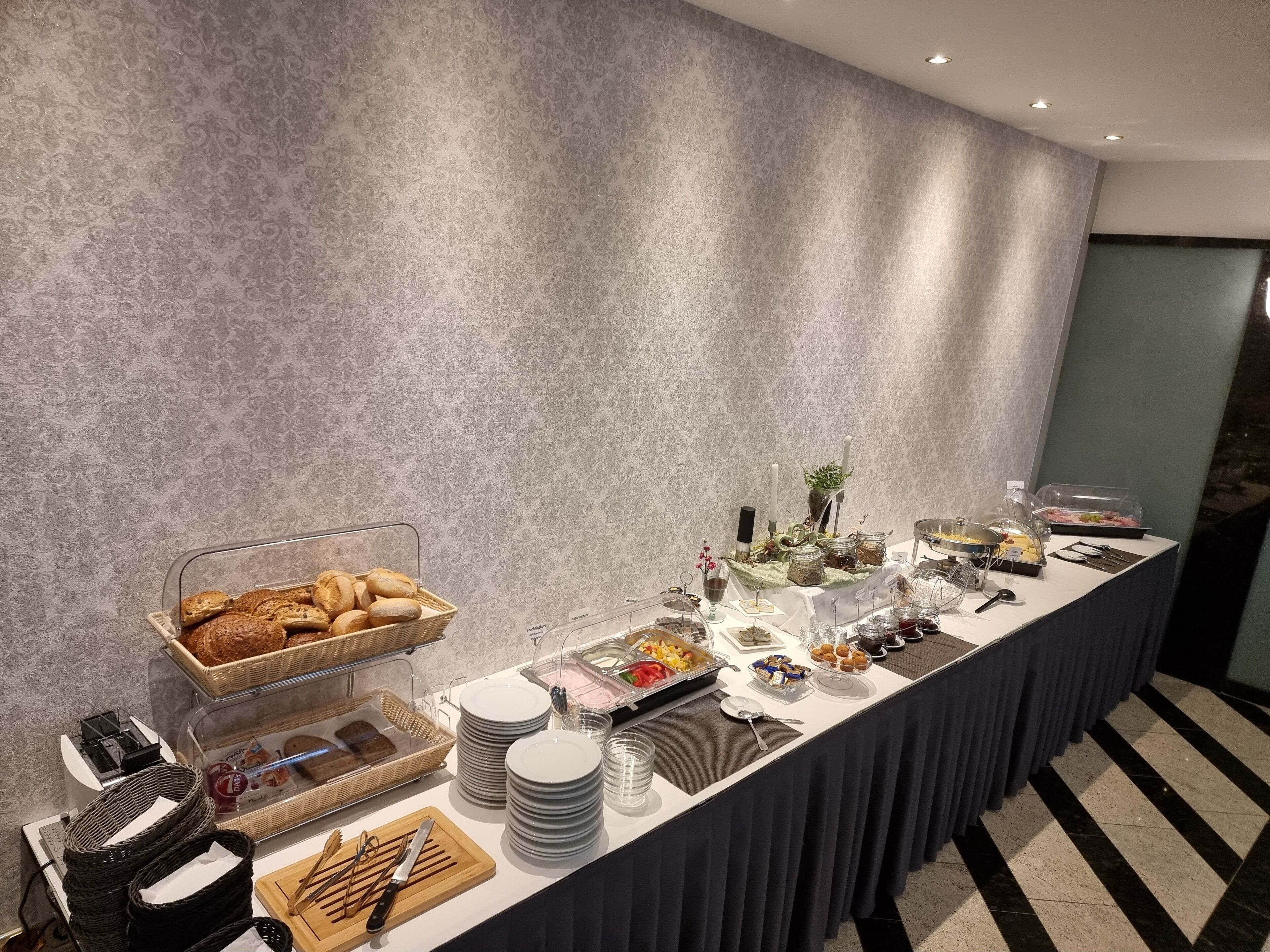 A buffet setup with various food items, including bread, fruits, and desserts, arranged on a long table with a patterned wall in the background.