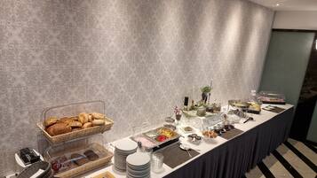 A buffet setup with various food items, including bread, fruits, and desserts, arranged on a long table with a patterned wall in the background.