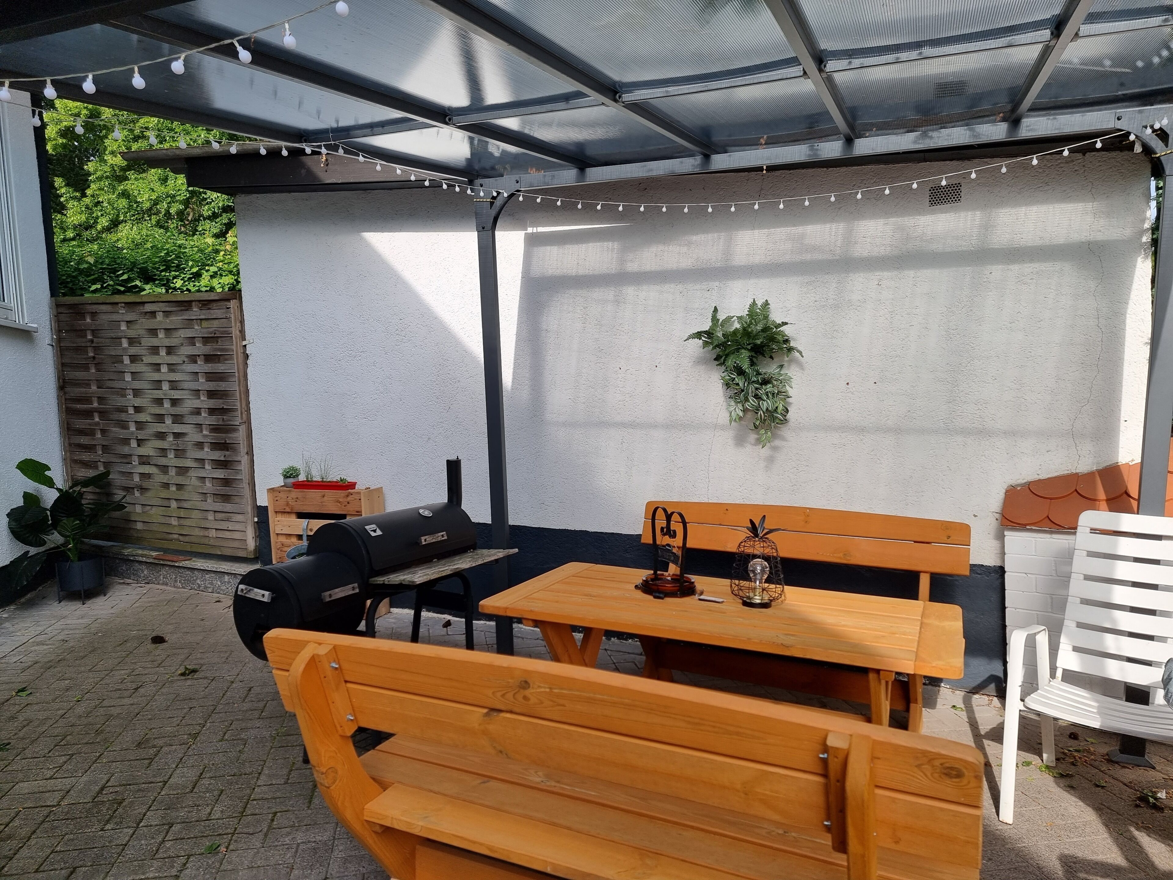 Ein überdachter Patio-Bereich mit Holztisch und Bänken, einem Grill und Lichterketten.