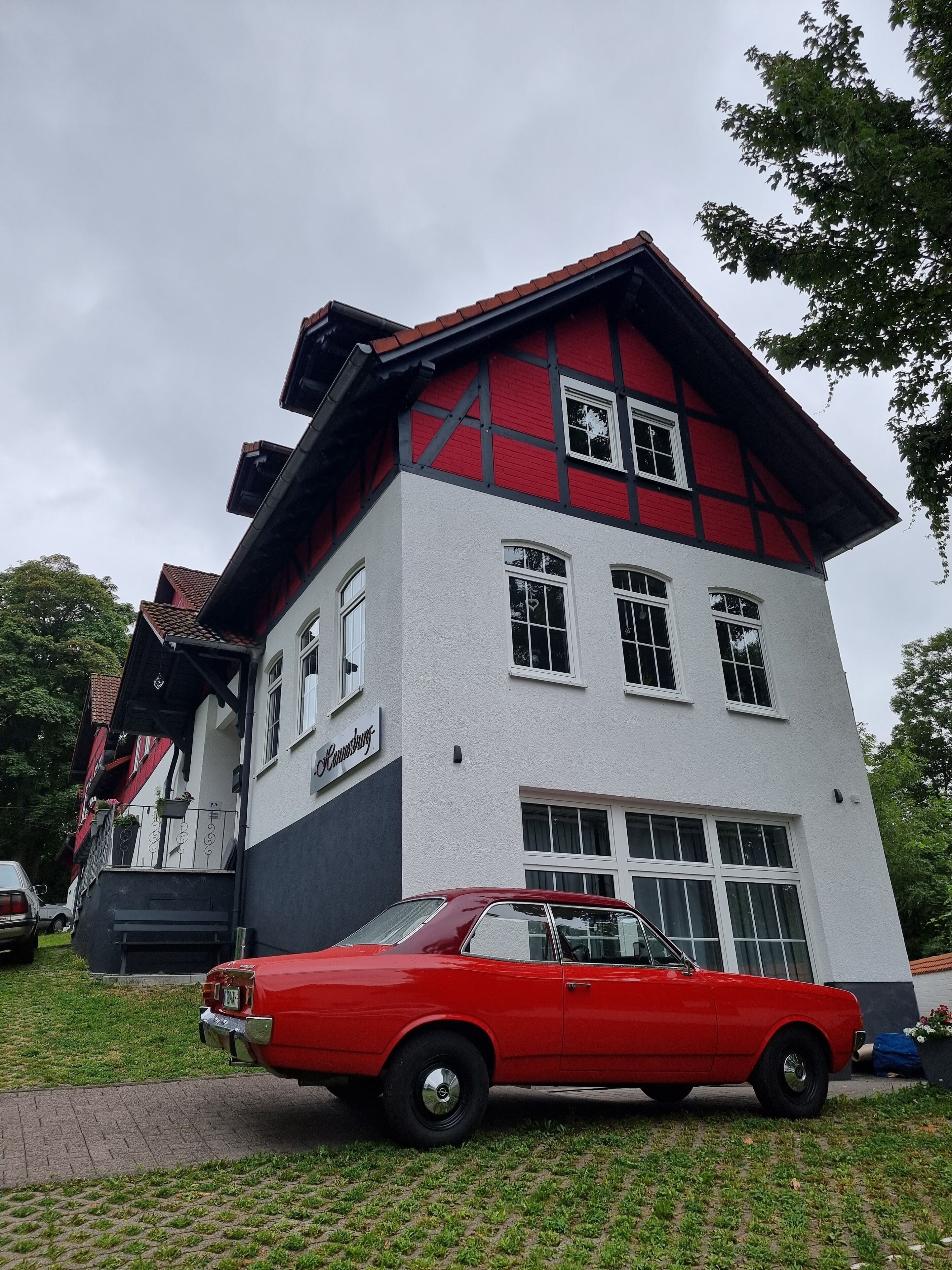 A two-story building with a red and white facade, a visible sign reading "Kaufhaus," and a red vintage car parked in front.