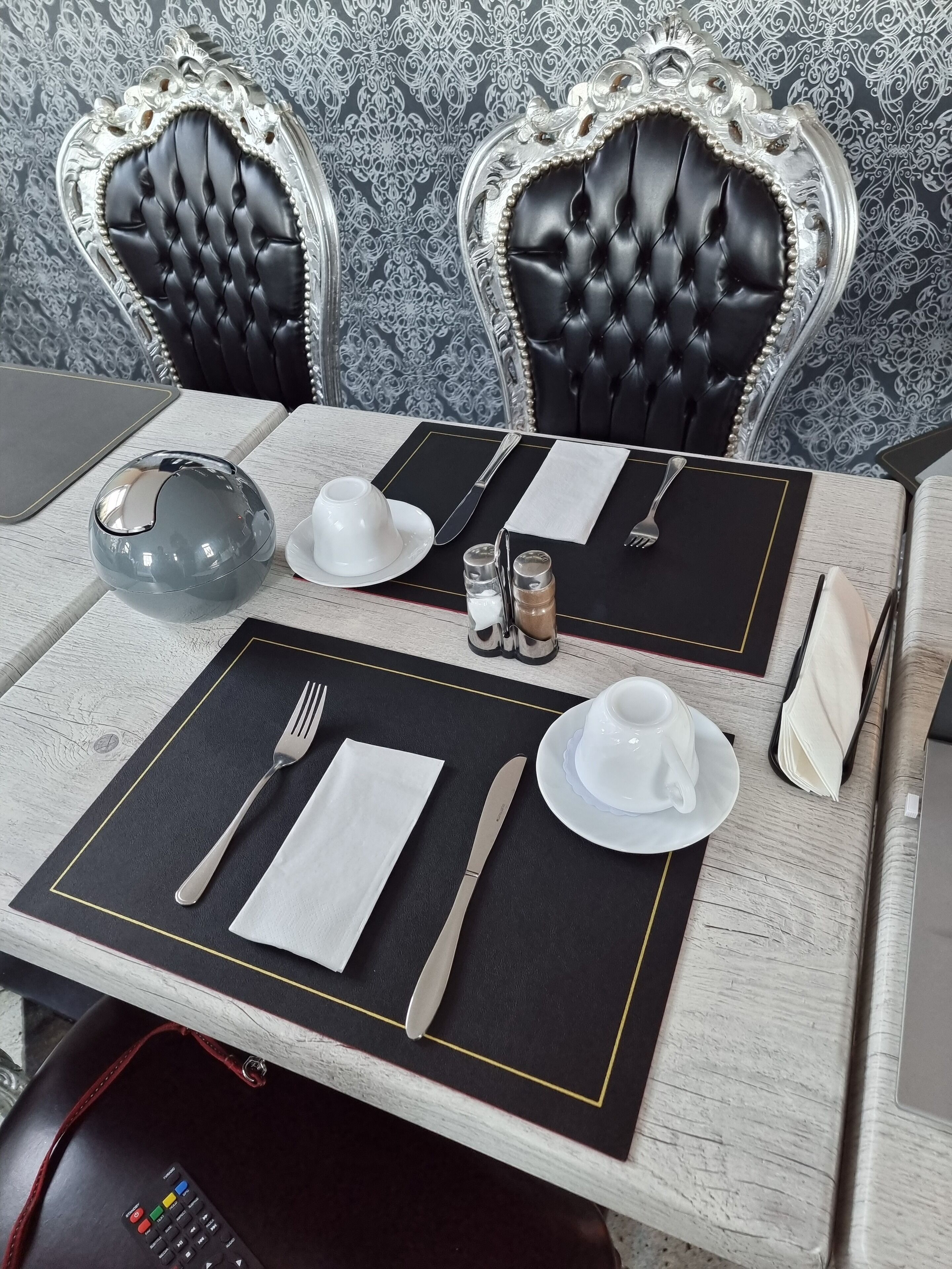 Ein Esstisch ist gedeckt mit einer schwarzen Tischmatte, einer weißen Serviette, Besteck und einer weißen Teetasse.