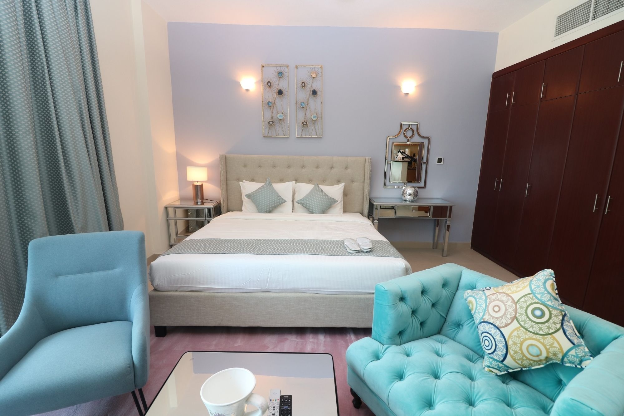 Une chambre d’hôtel comprenant un lit, une table de chevet, une commode, un miroir, un fauteuil bleu, un canapé turquoise et une table basse.
