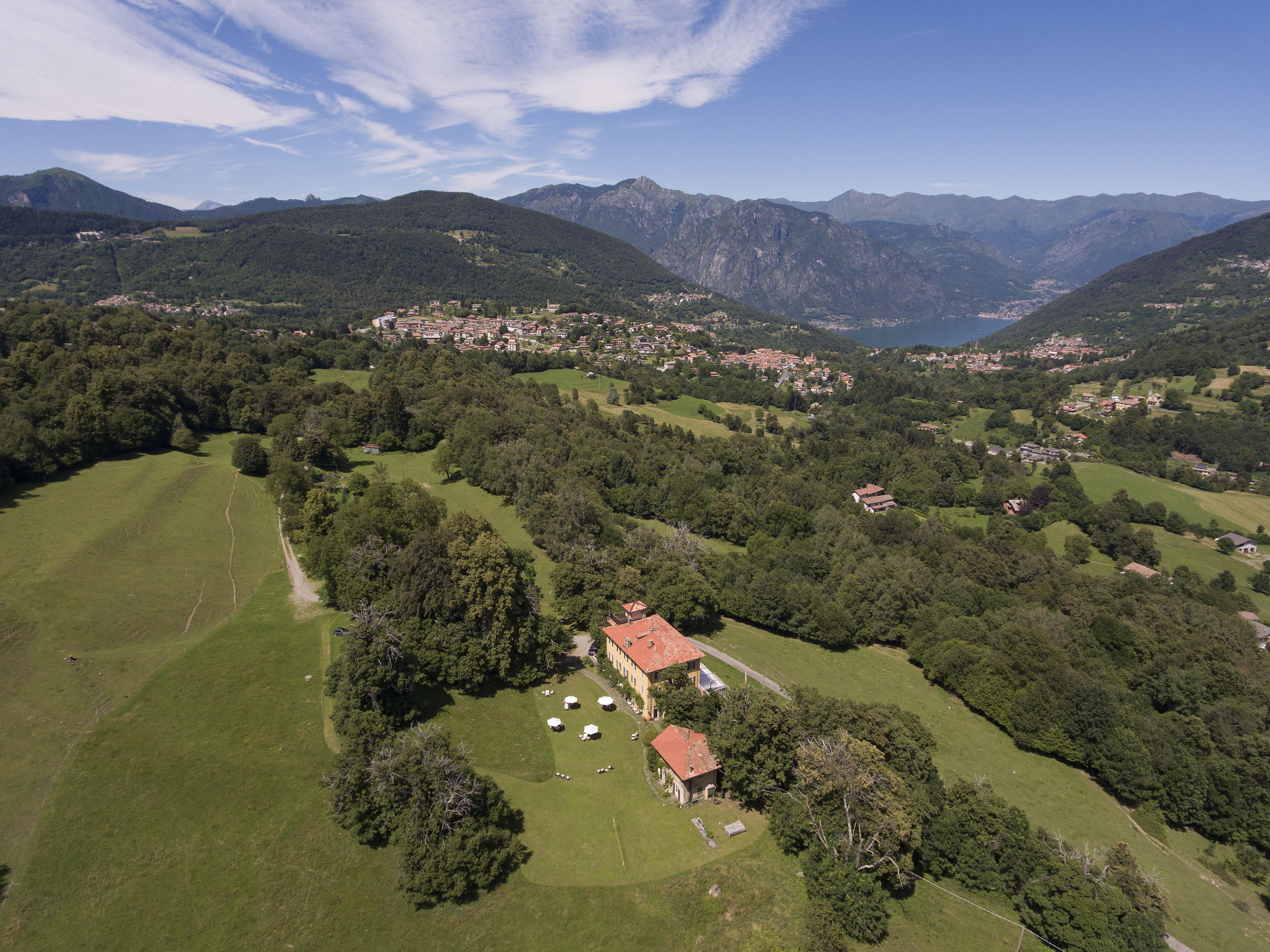 An aerial view of a rural area with a large house surrounded by green fields and trees, with mountains in the background.