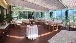 Un'area pranzo esterna coperta con tavoli e sedie, circondata da ampie finestre che offrono vista sul verde.