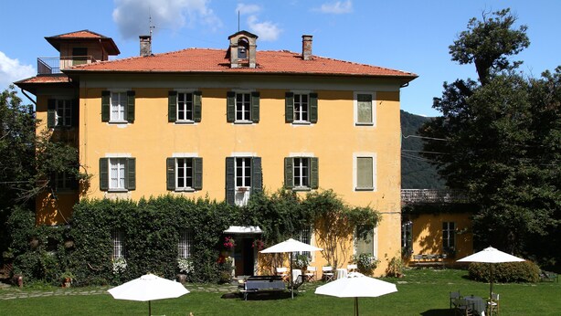 Un edificio giallo con persiane verdi, un tetto rosso e un giardino con posti a sedere all'aperto.