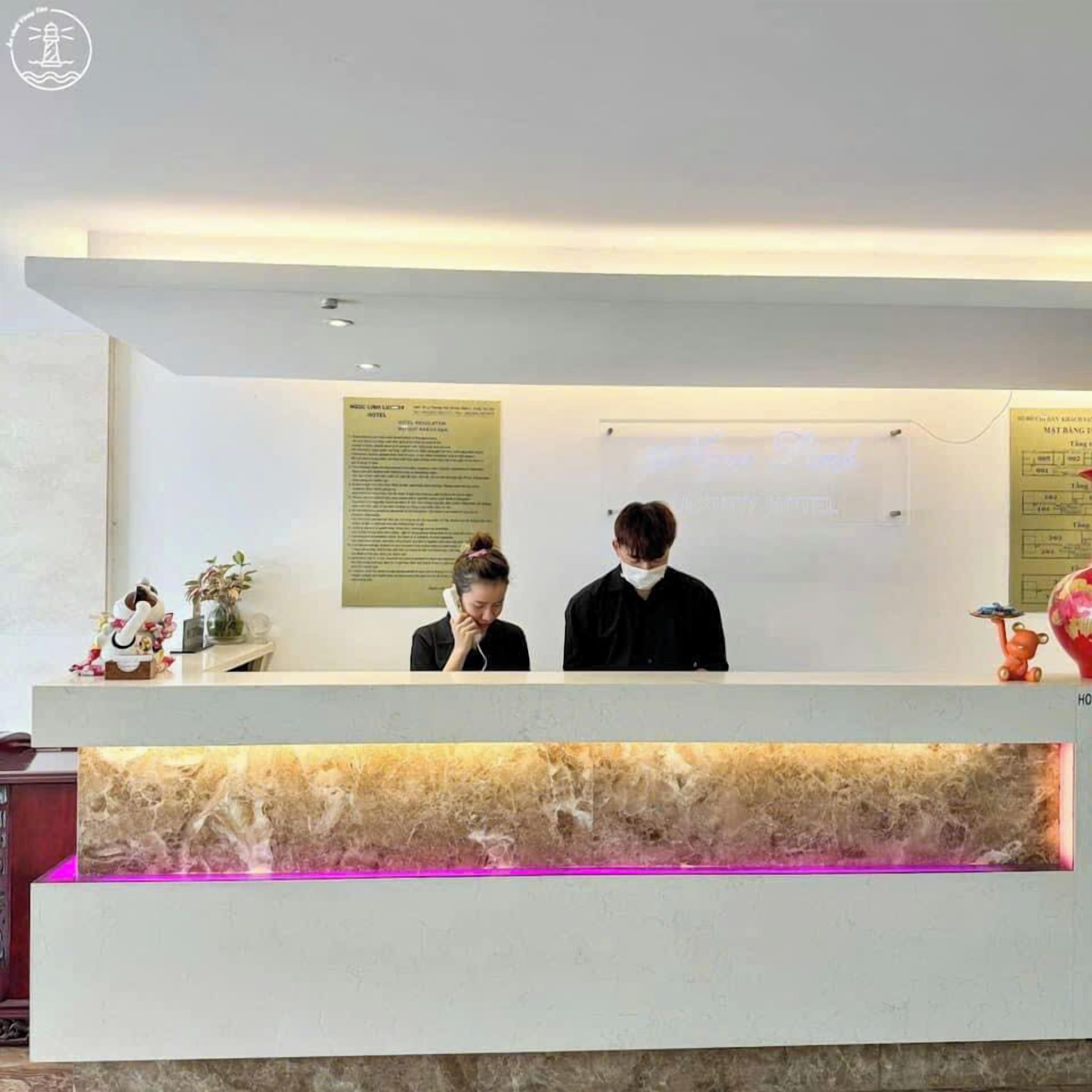Balcão de recepção de hotel com dois funcionários, um balcão de mármore e cartazes informativos na parede.