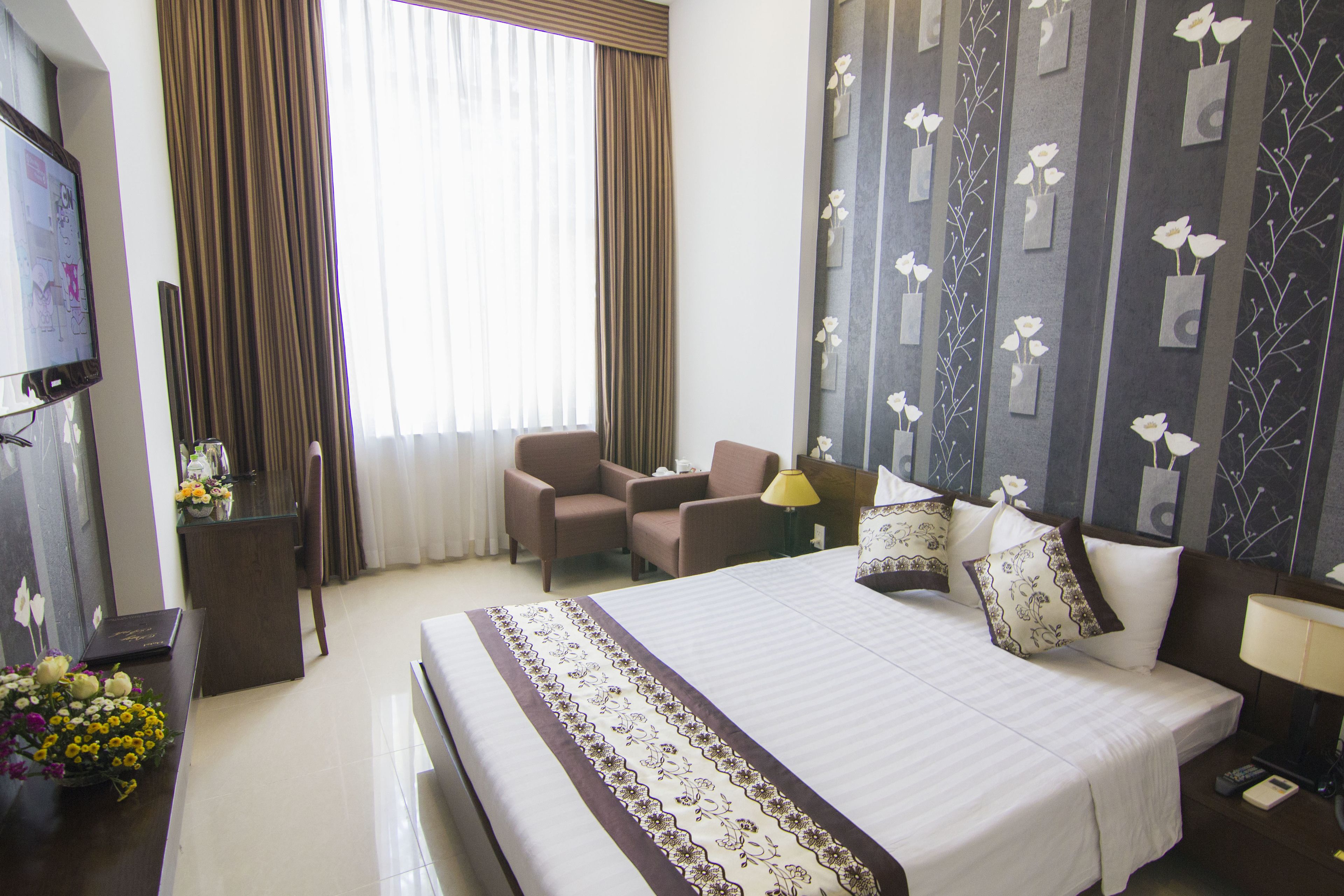 A hotel room with a large bed, two armchairs, a desk with a computer, and a TV.